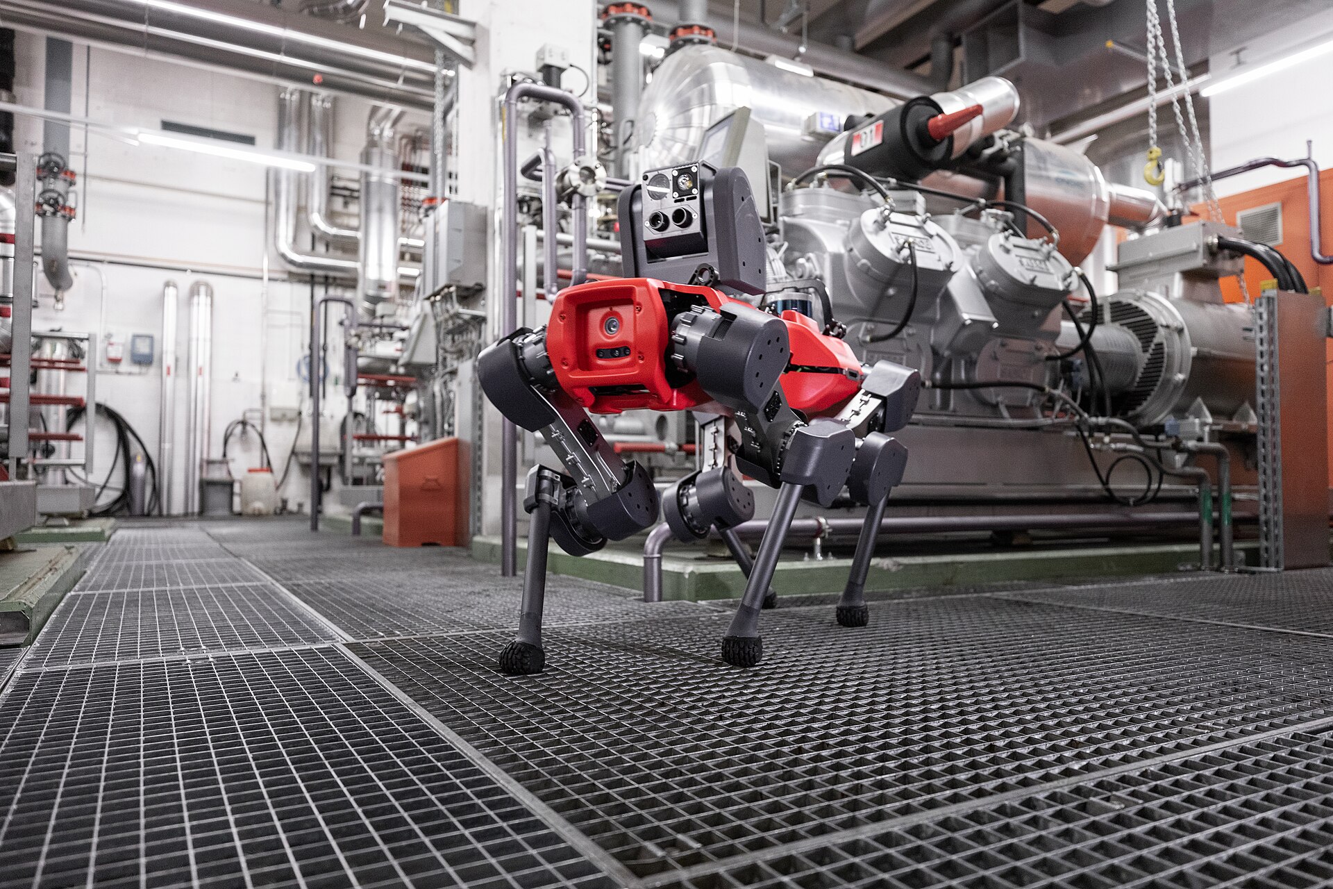 A red and grey ANYmal quadruped robot stands on a metal grate walkway inside an industrial facility, surrounded by pipes and machinery.