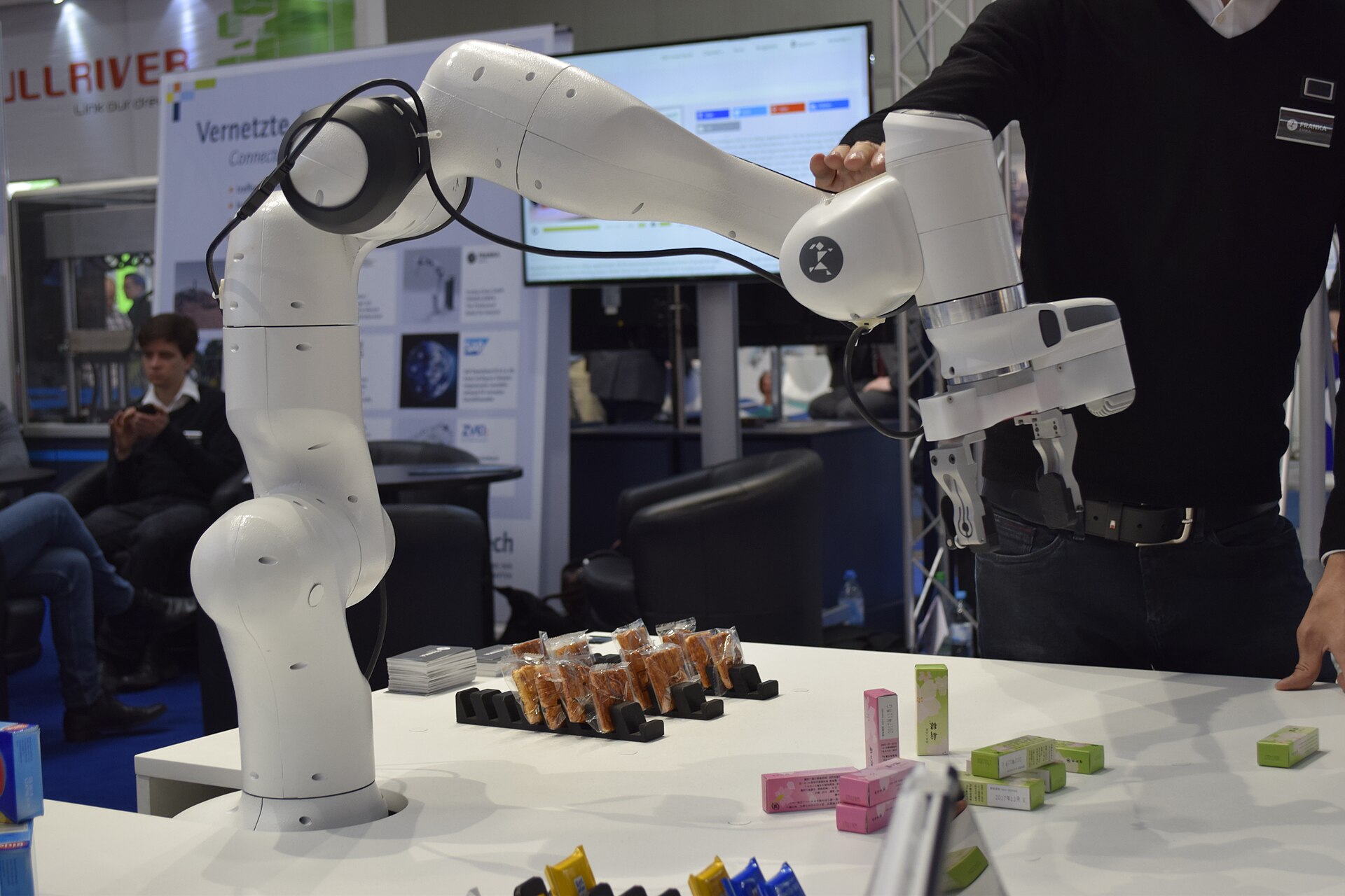 A white seven-axis Franka Emika Panda robot arm mounted on a demonstration table at CeBIT 2017, guided by hand above trays of small parts.