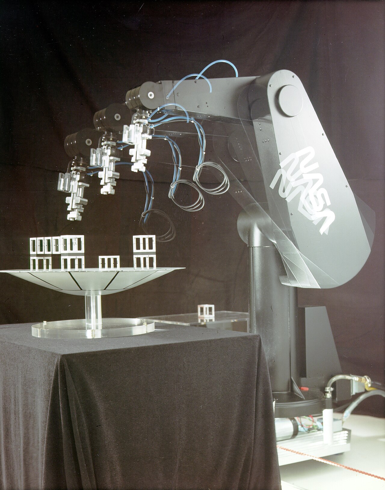 A stroboscopic NASA photograph shows a PUMA 560 robot arm in several successive positions as it moves small parts between fixtures on a table.