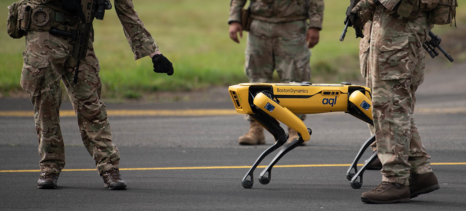 A yellow Boston Dynamics Spot quadruped robot walks across an airfield tarmac alongside Royal Air Force airmen in camouflage uniform.